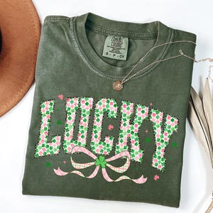 May include: An olive green t-shirt with the word "LUCKY" in a floral pattern of green shamrocks and pink flowers. A pink and green plaid bow is below the word. A gold necklace with a pendant is on the shirt.