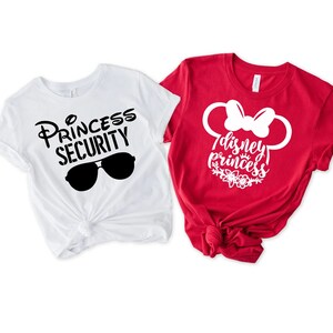 May include: Two white t-shirts with black text and graphics. One shirt says "Princess Security" with sunglasses. The other shirt is red with white text and a white graphic of Mickey Mouse ears with a bow. The text says "Disney Princess".
