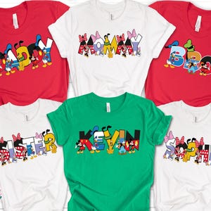 May include: A collection of colorful t-shirts featuring Disney characters and names. The shirts are red, white, and green, with names like "Daddy," "Mommy," "Bro," "Sister," "Kevin," and "Sophia" in a playful font, perfect for family or group wear.