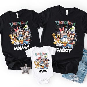 May include: Black t-shirts and a white onesie featuring a colorful graphic of Disney characters. The shirts have "Disneyland 2026" and "Mommy" or "Daddy" printed on them. The onesie has "Disneyland 2026" and "Name" printed on it.
