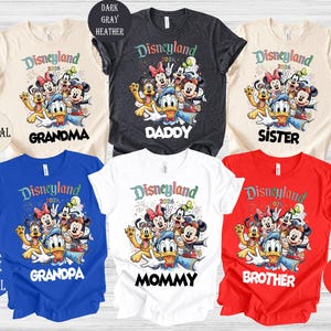 May include: A collection of colorful t-shirts featuring a Disneyland 2026 graphic with various Disney characters. The shirts are in natural, dark gray, blue, white, and red, with text for Grandma, Daddy, Sister, Grandpa, Mommy, and Brother.