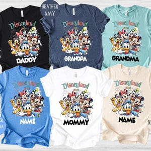 May include: Assorted-color t-shirts with a Disneyland 2026 graphic featuring Mickey Mouse, Minnie Mouse, Donald Duck, and other characters. The shirts are available in black, navy, dusty blue, heather blue, white, and natural colors, with text for family members.