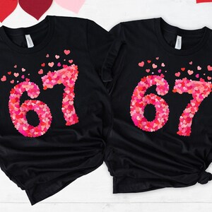 May include: Two black t-shirts with the number "67" in pink and red heart shapes. The hearts are arranged to form the numbers, with some hearts scattered around them. The shirts are made of a soft material.