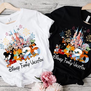 Peut inclure: T-shirts blancs et noirs avec un graphisme color&eacute; repr&eacute;sentant des personnages Disney, un ch&acirc;teau et le texte "2020 Disney Family Vacation". Les chemises sont suspendues &agrave; des cintres en bois.