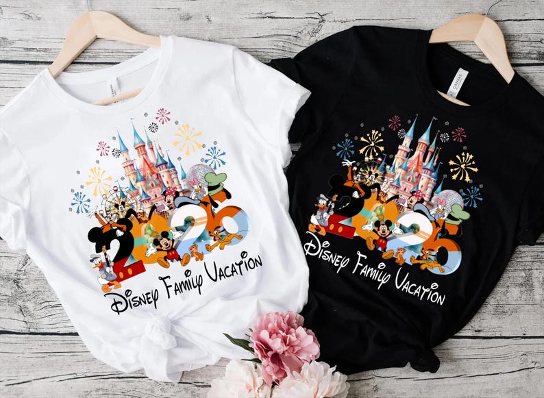 Peut inclure: T-shirts blancs et noirs avec un graphisme color&eacute; repr&eacute;sentant des personnages Disney, un ch&acirc;teau et le texte "2020 Disney Family Vacation". Les chemises sont suspendues &agrave; des cintres en bois.