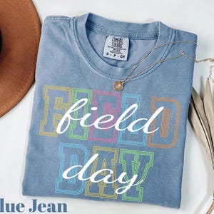 May include: A blue jean colored t-shirt with the words "Field Day" in a colorful, outlined font. The shirt has a crew neck and short sleeves. A gold necklace with a pendant is draped across the shirt.