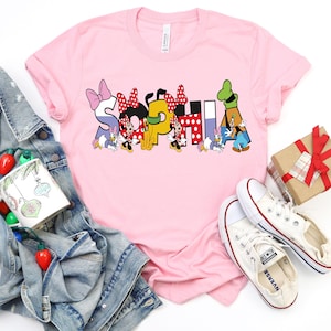 May include: A light pink t-shirt with the name "SOPHIA" in colorful Disney-themed letters. The letters are decorated with Minnie Mouse, Daisy Duck, and Donald Duck characters. White sneakers and a denim jacket are also in the image.