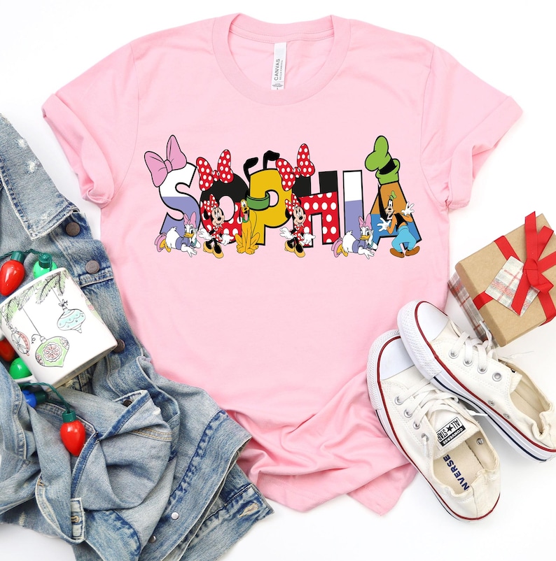 May include: A light pink t-shirt with the name "SOPHIA" in colorful Disney-themed letters. The letters are decorated with Minnie Mouse, Daisy Duck, and Donald Duck characters. White sneakers and a denim jacket are also in the image.