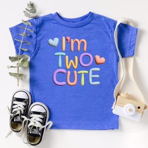 Puede incluir: Una camiseta azul para ni&ntilde;os peque&ntilde;os con el texto "I'm Two Cute" en colores arco&iacute;ris y corazones.