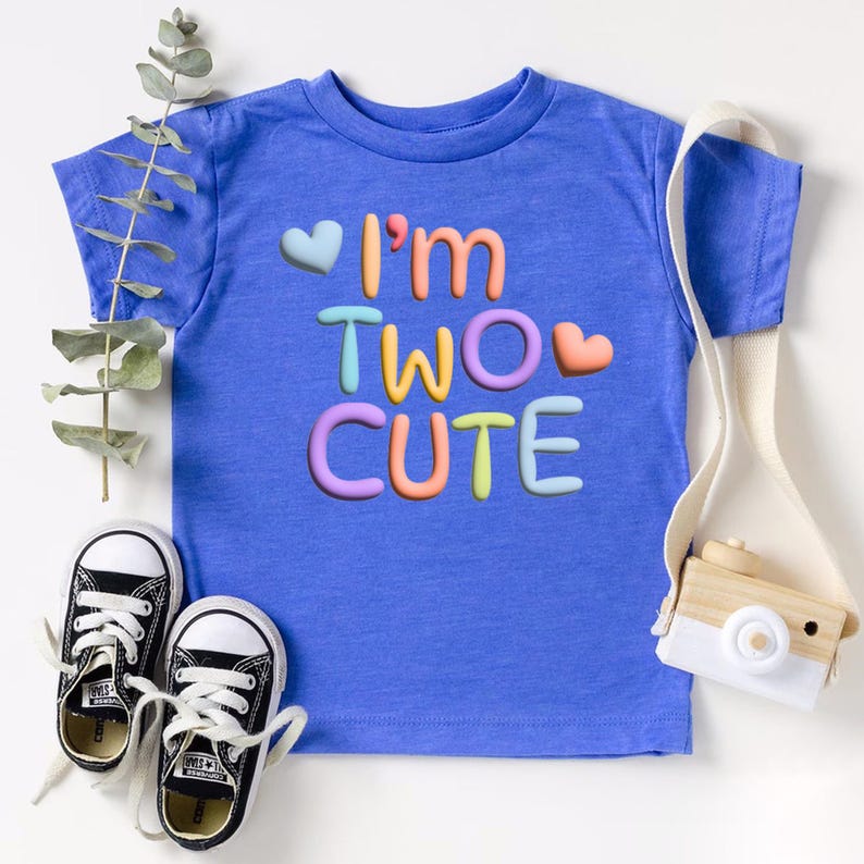 Puede incluir: Una camiseta azul para ni&ntilde;os peque&ntilde;os con el texto "I'm Two Cute" en colores arco&iacute;ris y corazones.