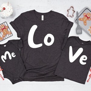 Puede incluir: Un conjunto de tres camisetas grises oscuras con las palabras "Yo", "Amor" y "T&uacute;" en blanco. Las camisetas est&aacute;n dispuestas sobre una superficie blanca con galletas de jengibre y otras decoraciones navide&ntilde;as.