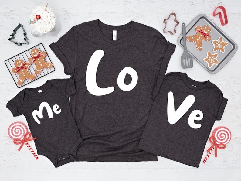 Puede incluir: Un conjunto de tres camisetas grises oscuras con las palabras "Yo", "Amor" y "T&uacute;" en blanco. Las camisetas est&aacute;n dispuestas sobre una superficie blanca con galletas de jengibre y otras decoraciones navide&ntilde;as.