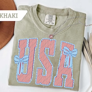Może przedstawiać: Khaki koszulka z napisem "USA" w czerwono-białe paski, obramowana na niebiesko z niebieskimi kokardkami. Srebrny naszyjnik z wisiorkiem spoczywa na koszulce. Koszulka jest oznaczona jako "Comfort Colors".