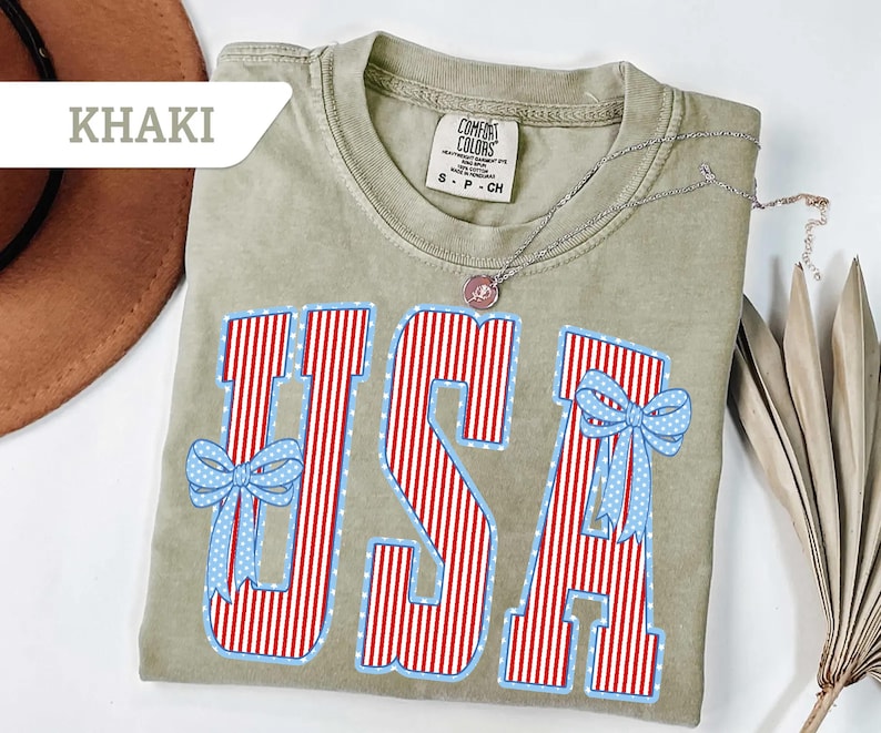 Może przedstawiać: Khaki koszulka z napisem "USA" w czerwono-białe paski, obramowana na niebiesko z niebieskimi kokardkami. Srebrny naszyjnik z wisiorkiem spoczywa na koszulce. Koszulka jest oznaczona jako "Comfort Colors".