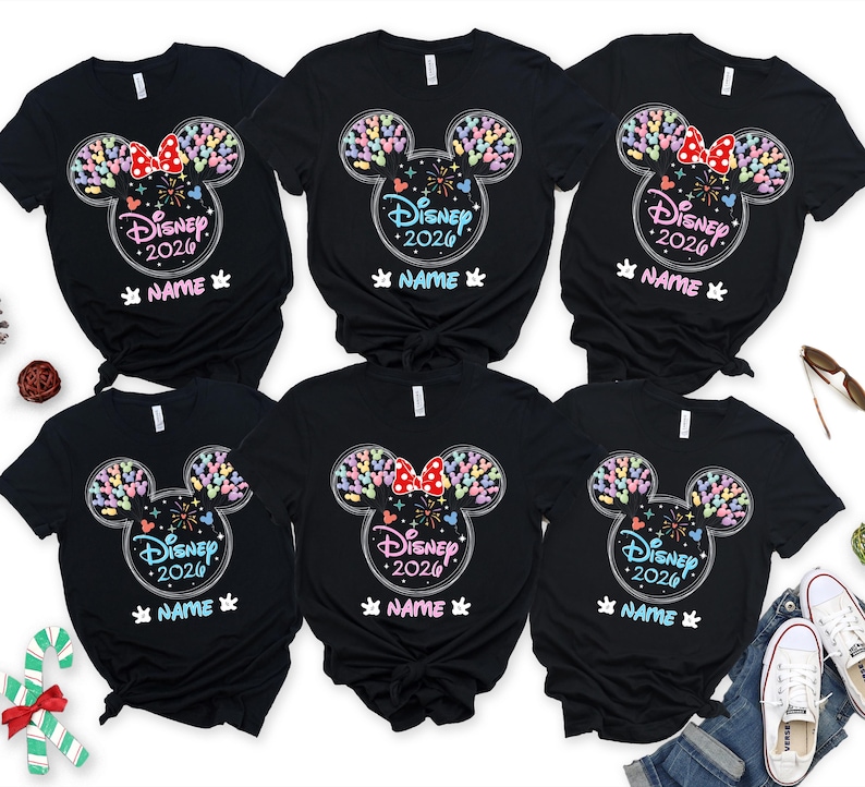 Puede incluir: Camisetas negras con un dise&ntilde;o de Disney. El dise&ntilde;o presenta una forma de cabeza de Mickey Mouse llena de formas coloridas, el texto "Disney 2026" y un espacio para un nombre. Algunas camisetas tienen un lazo rojo.