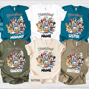 May include: A collection of colorful Disneyland 2026 graphic t-shirts in various colors, including teal, white, olive, and natural. Each shirt features a Disney character design and text such as "Mommy," "Sister," "Daddy," "Brother," and "Name."