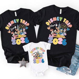 K&ouml;nnte beinhalten: Schwarze T-Shirts und ein wei&szlig;er Body mit einem farbenfrohen Disney Trip 2026 Design. Die Shirts zeigen Mickey und Minnie Mouse, Donald und Daisy Duck und andere Disney-Figuren. Der Body hat das gleiche Design.