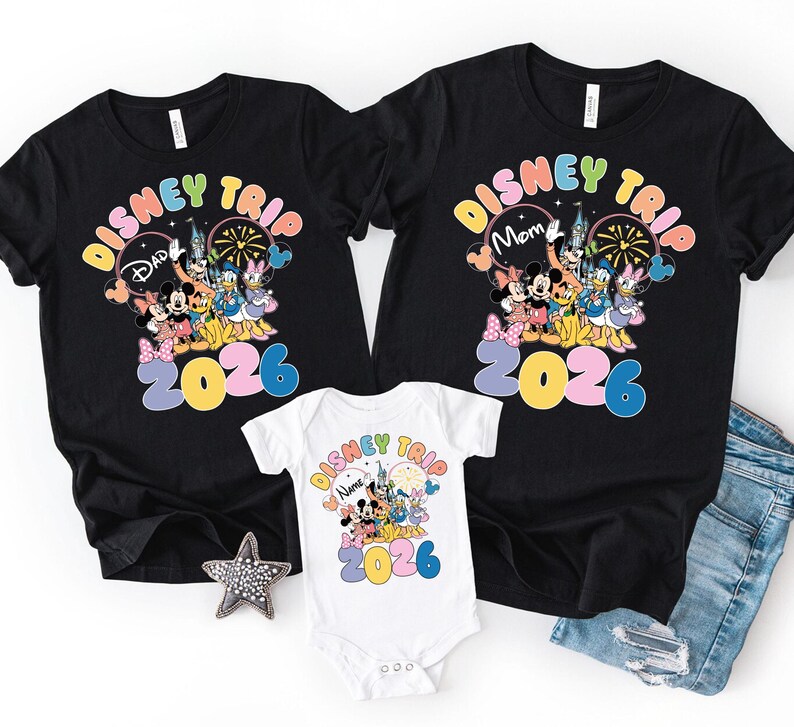 K&ouml;nnte beinhalten: Schwarze T-Shirts und ein wei&szlig;er Body mit einem farbenfrohen Disney Trip 2026 Design. Die Shirts zeigen Mickey und Minnie Mouse, Donald und Daisy Duck und andere Disney-Figuren. Der Body hat das gleiche Design.
