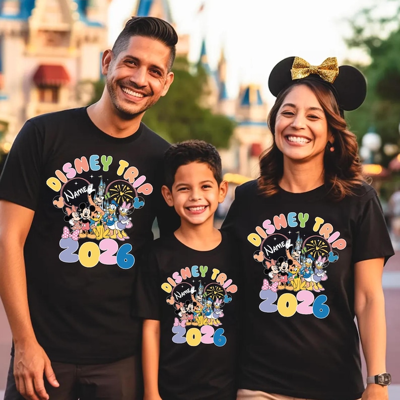 K&ouml;nnte beinhalten: Drei Personen tragen passende schwarze T-Shirts mit einer farbenfrohen Disney-Grafik. Die Shirts tragen die Aufschrift "Disney Trip" mit Platz f&uuml;r einen Namen und dem Jahr "2026". Die Grafik zeigt Micky Maus, Minnie Maus und andere Disney-Figuren.
