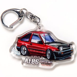 Op de afbeelding: Rode en zwarte Toyota AE86 Trueno GT-Apex sleutelhanger met een zilveren sleutelring. De sleutelhanger heeft een cartoonillustratie van de auto met de tekst "ハチロク AE86 TRUENO GT-APEX TWIN CAM 16" eronder.