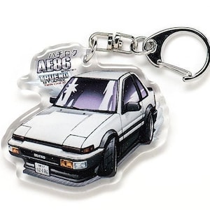 Op de afbeelding: Witte Toyota AE86 Trueno Twin Cam Apex sleutelhanger met een zilveren sleutelring. De sleutelhanger heeft een cartoonillustratie van de auto met de tekst "ハチロク AE86 TRUENO TWIN CAM APEX" aan de voorkant.