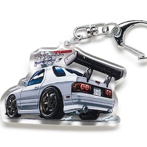 Op de afbeelding: Een heldere acryl sleutelhanger met een witte sportwagen met een grote achtervleugel. De auto heeft bruine wielen en een kentekenplaat. De sleutelhanger heeft een zilveren metalen sluiting en een splitring. De tekst "TURBO SAVANNA RX-7" is zichtbaar.