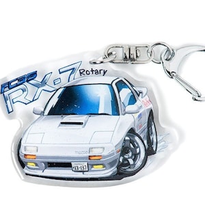 Op de afbeelding: Een witte Mazda RX-7 sleutelhanger met een blauwe en witte cartoon-illustratie van de auto. De sleutelhanger heeft een zilveren metalen ring en een transparante acryl achtergrond. De tekst "Rotary" is op de sleutelhanger gedrukt.
