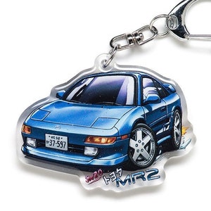 May include: A blue and white keychain featuring a cartoon illustration of a Toyota MR2 sports car with the text "SW20 トヨタ MR2" on the bottom. The keychain has a silver metal ring.
