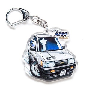 May include: A white Toyota AE85 Levin keychain featuring a blue and white cartoon illustration of the car. The keychain has a silver metal ring.