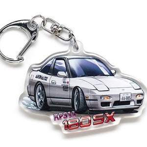 Op de afbeelding: Een witte Nissan 180SX sleutelhanger met een zilveren sleutelring. De sleutelhanger heeft een cartoonillustratie van de auto met de tekst "RPS13 180SX" en "AKINA SPEED" aan de zijkant. De auto heeft een Japans kenteken met de tekst "11-305".