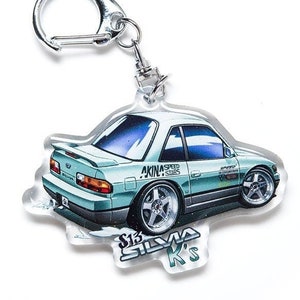 Op de afbeelding: Een lichtblauwe en zilveren sleutelhanger met een cartoonillustratie van een Nissan S13 Silvia met de tekst "AKINA SPEED STARS" en "S13 SILVIA K'S" aan de zijkant.