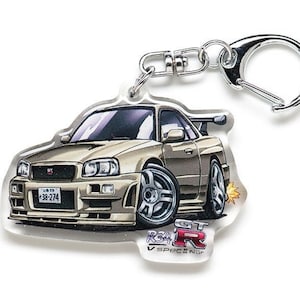 Op de afbeelding: Een zilveren sleutelhanger met een beige Nissan Skyline GT-R R34 V-Spec II Nur autobedel. De auto heeft een zwart kenteken met de tekst "33-274".