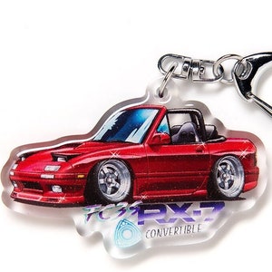 Puede incluir: Un llavero de coche descapotable rojo con detalles plateados. El llavero presenta una ilustración estilizada de un coche deportivo con el texto "RX-7 Convertible". El llavero está unido a un anillo de metal plateado.