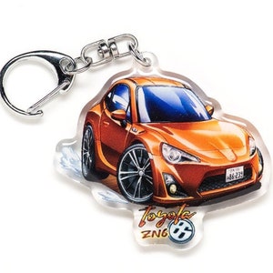 Puede incluir: Llavero Toyota 86 naranja con un llavero plateado. El llavero presenta una ilustración de dibujos animados del coche con el texto "Toyota 86 ZN6" en la parte inferior.
