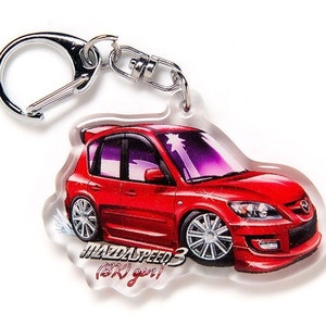 Op de afbeelding: Rode MazdaSpeed 3 sleutelhanger met een zilveren sleutelring. De sleutelhanger heeft een cartoonillustratie van een rode MazdaSpeed 3 met de tekst "MazdaSpeed 3 (RX-8 gen)" onder de auto.