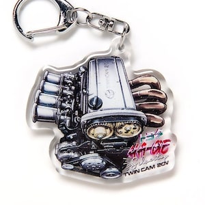 Op de afbeelding: Een zilveren en zwarte auto-motor sleutelhanger met een rode en witte afbeelding met de tekst "4A-GE Silvertop Twin Cam 20V".