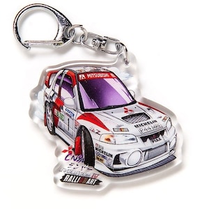 Könnte beinhalten: Ein weißer Mitsubishi Lancer Evolution Rallye-Auto-Schlüsselanhänger mit roten und schwarzen Akzenten. Der Schlüsselanhänger trägt den Text "CN9A EVOLUTION IV RALLY ART" und "MICHELIN P44 MRC".