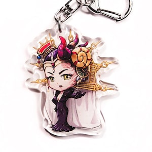 May include: A clear acrylic keychain featuring a cartoon character with purple hair, a crown, and horns. The character is wearing a black dress and a white cape. The keychain is attached to a silver metal ring.