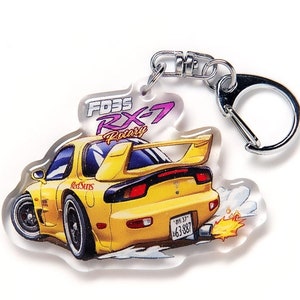 Op de afbeelding: Een gele Mazda RX-7 FD3S sleutelhanger van helder acryl met een rode en witte RedSuns-sticker op de achterkant. De sleutelhanger heeft een cartoonachtige illustratie van de auto met een vlam uit de uitlaat.