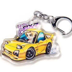 May include: A yellow RX-7 car keychain with a cartoon driver in the driver's seat. The keychain has a silver metal ring.