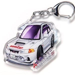 Può includere: Un portachiavi in acrilico trasparente con un'auto bianca con vetri oscurati viola. L'auto ha la scritta "monster" in rosso sul cofano e testo giapponese. Il portachiavi ha un fermaglio argentato.