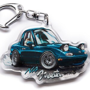 Op de afbeelding: Een blauwe en zilveren sleutelhanger met een cartoonillustratie van een Mazda Miata met de tekst "NA Miata MX-5".