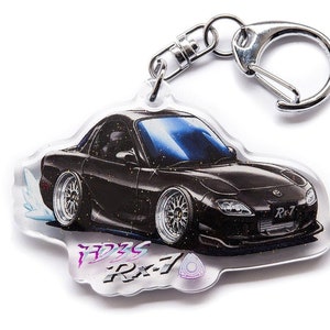 Op de afbeelding: Een zwarte Mazda RX-7 sleutelhanger met een zilveren sleutelring. De sleutelhanger is gemaakt van helder acryl en heeft een gedetailleerde illustratie van de auto met een wit rookstreepje erachter. De tekst "FD3S RX-7" is in een gestileerd lettertype op de sleutelhanger gedrukt.