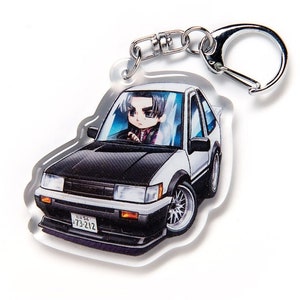 May include: White and black anime-style keychain featuring a cartoon character driving a white car with a black hood. The car has a Japanese number plate that reads "73-212".