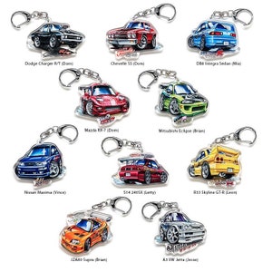 Op de afbeelding: Tien verschillende autosleutelhanger met verschillende merken en modellen van auto's. Elke sleutelhanger is een transparante acryl hanger met een kleurrijke cartoon-illustratie van een auto. De auto's omvatten een Dodge Charger, een Chevelle SS, een Integra Sedan, een Mazda RX-7, een Mitsubishi Eclipse, een Nissan Maxima, een S14 240SX, een R33 Skyline GT-R, een JZA80 Supra en een VW Jetta.