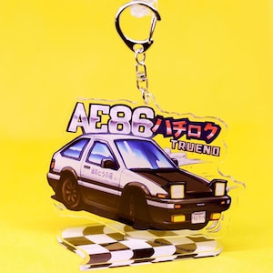 Op de afbeelding: Een witte en zwarte anime-stijl auto sleutelhanger met de tekst "AE86 ハチロク TRUENO" en een geruite racebaan basis.