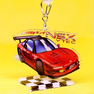 Op de afbeelding: Een rode Honda NSX auto-sleutelhanger met een zwarte spoiler en een basis van een geblokte vlag. De sleutelhanger heeft de tekst "NSX VTEC" en "ボック" in het Japans.