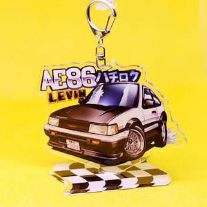 Op de afbeelding: Een heldere acryl sleutelhanger met een wit en zwarte auto met de tekst "AE86 LEVIN" en Japanse karakters. De sleutelhanger heeft een zilveren metalen sluiting en een geblokte basis.