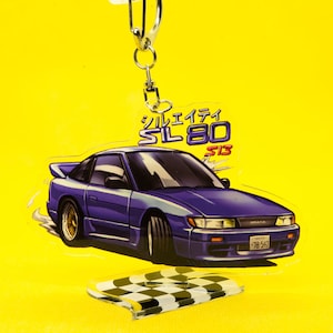 Op de afbeelding: Een blauwe en witte auto sleutelhanger met een geruite basis. De sleutelhanger heeft een blauwe auto met een wit interieur en de tekst "シルエイティ SIL80 S13" in het Japans.