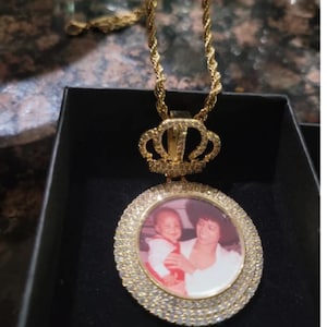 Puede incluir: Collar con colgante dorado con una foto circular rodeada de peque&ntilde;as piedras transparentes. El colgante est&aacute; rematado con un dise&ntilde;o de corona y cuelga de una cadena de cuerda. La foto muestra a una madre y su hijo.